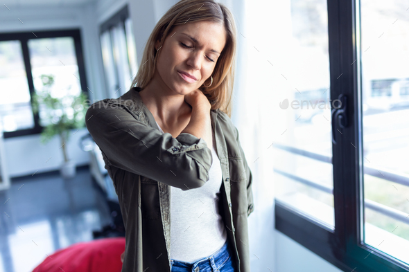 Tired young woman with neck and back pain standing in the living room at home. - Stock Photo - Images