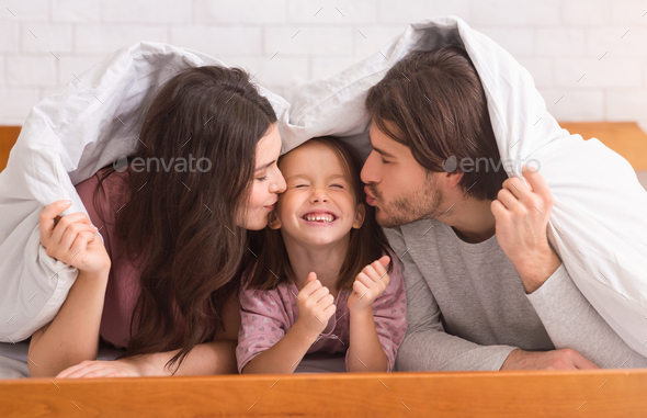 Parents kissing their daughter while hiding under blanket, having fun together