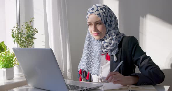 Arab young woman student writes in a notebook and watches a lesson on a laptop. alt