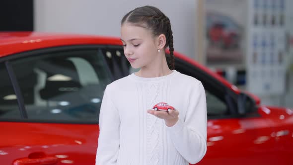 Portrait of Beautiful Charming Caucasian Girl Standing in Car Dealership with Toy Red Automobile alt