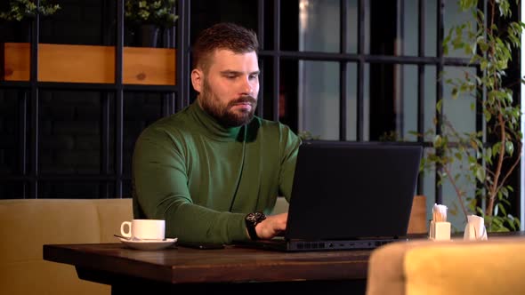 Young Attractive Bearded Man Confidently Working on Laptop During Coffee Break in Cafe