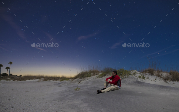 A man sits alone on a beach at night with stars above.