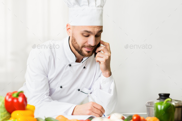 Male Chef Talking On Cellphone Taking Notes Standing In Kitchen