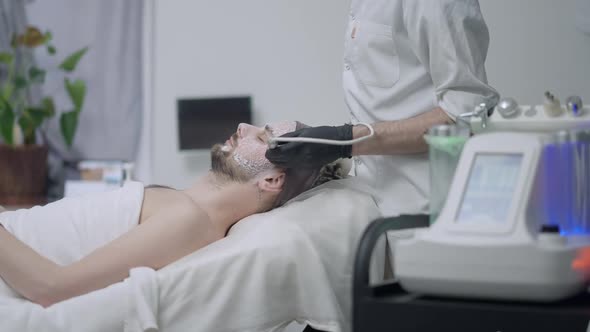 Side View of Relaxed Handsome Man Lying in Beauty Salon As Unrecognizable Beautician Using LED Light alt