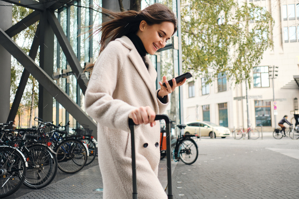 Young businesswoman in coat happily using cellphone walking through street with suitcase