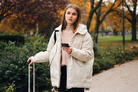Attractive casual girl in down jacket with cellphone thoughtfully looking away in city park