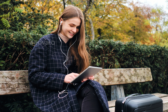 Pretty casual girl in earphones happily using tablet sitting with suitcase on bench in park