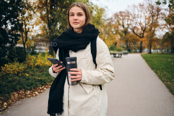 Pretty casual girl in down jacket walking around park alone with cellphone and thermo cup