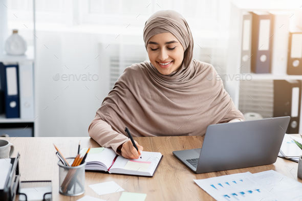 Smiling Islamic Businesswoman Using Laptop And Writing Notes, Managing Her Schedule