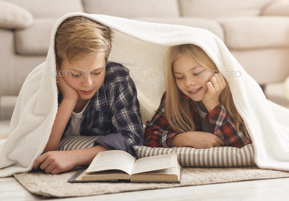 Brother And Sister Reading Book Covered With Blanket At Home