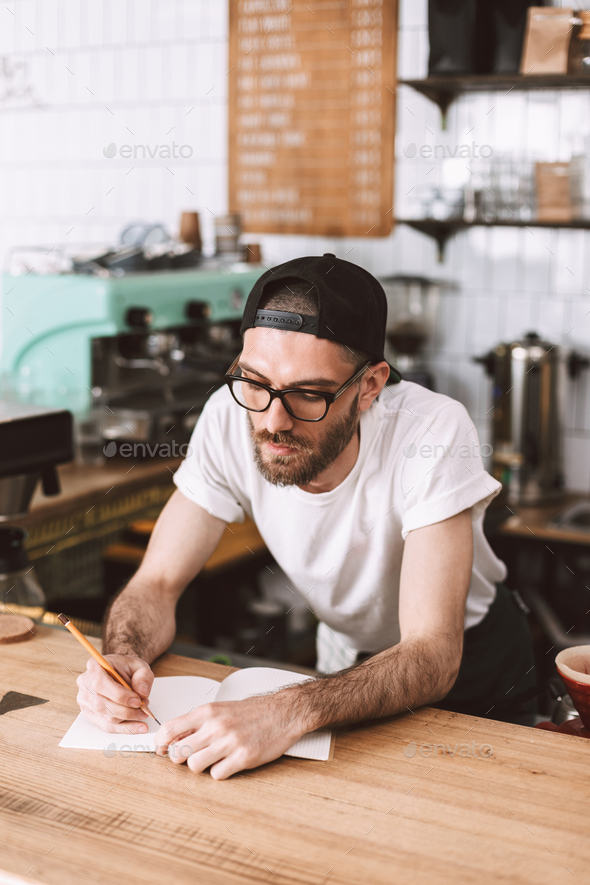 Pensive man in eyeglasses and cap standing behind bar counter writing in notepad working in cafe