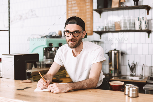 Smiling man in eyeglasses and cap standing behind bar counter with pencil and notepad
