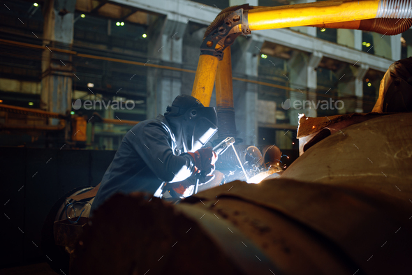Welder in mask works with metal pipe on factory