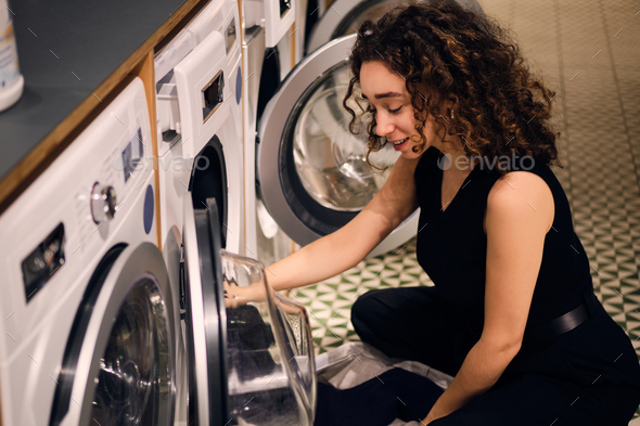 Beautiful dreamy girl loading clothes into washing machine in self-service laundry