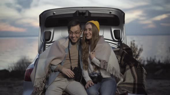 Positive Young Loving Couple Sitting in Car Trunk Using Binoculars alt