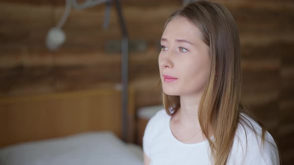 Young Beautiful Frustrated Woman Looking Out the Window in Hospital Ward Closing Eyes Sighing alt