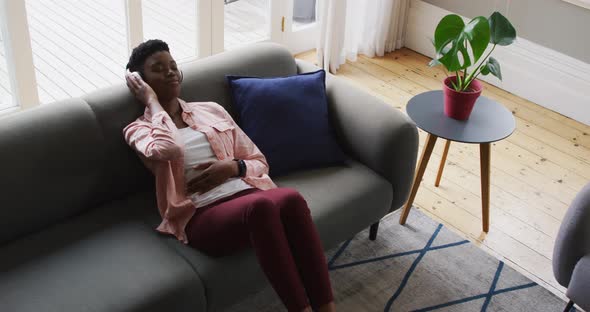 African american woman listening to music on headphones while lying on the couch at home alt