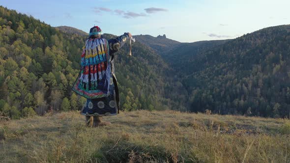 An Old Shaman Performs a Magic Ritual with the Help of a Bell Standing on the Top of a Mountain