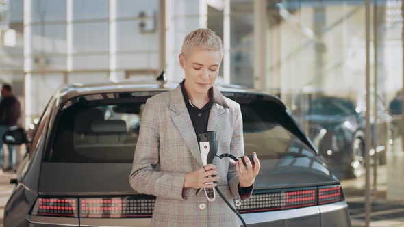 Blond Caucasian Middle Age Smiling Woman at an Electric Car Charging Station alt