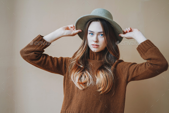 Young beautiful girl with blue eyes in felt hat and brown knitted sweater on beige background