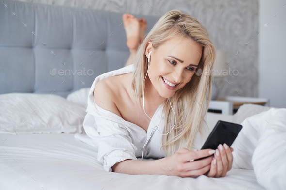 Adorable, smiling blond woman lying in white bed and using a smartphone - Stock Photo - Images