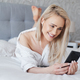 Adorable, smiling blond woman lying in white bed and using a smartphone - PhotoDune Item for Sale
