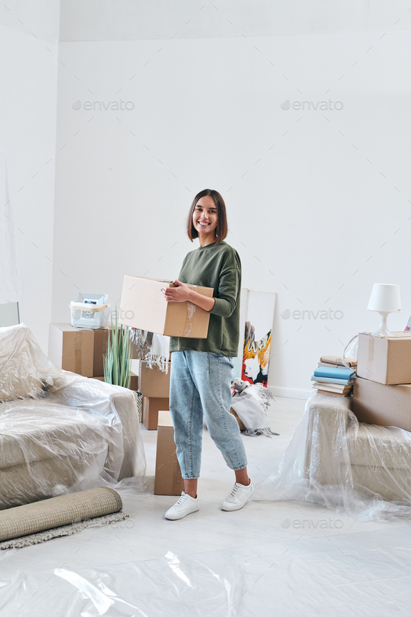 Young cheerful woman in casualwear carrying box while moving along room
