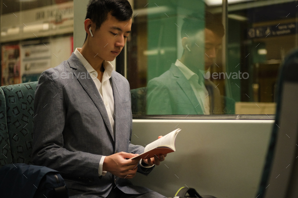 Young Asian businessman in wireless earphones intently reading book in subway train alone