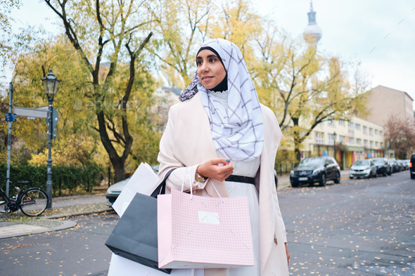 Young attractive Arabic woman in hijab dreamily walking with shopping bags on street
