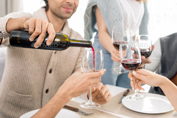 Young man pouring red wine from bottle into wineglasses of his friends