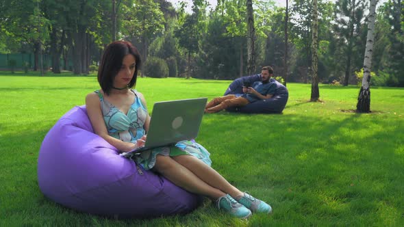 The Girl Brunette Sits in a Chair in the Park and Working on a Laptop alt