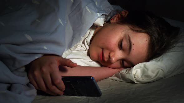 Little Girl Looking at The Smartphone Under The Blanket on Bed in a Dark Bedroom