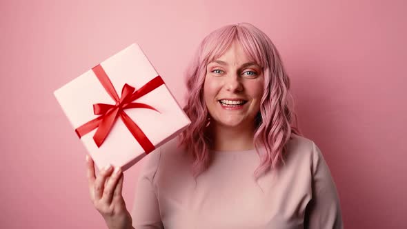 Happy Positive Young Caucasian Woman Showing Gift Box While Looking at Camera Isolated on Pink