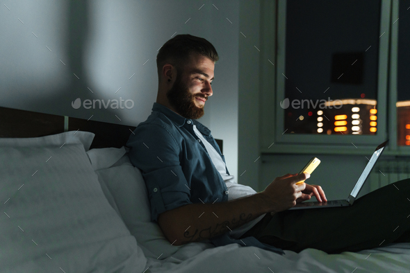 Smiling young bearded man laying on bed at home at night
