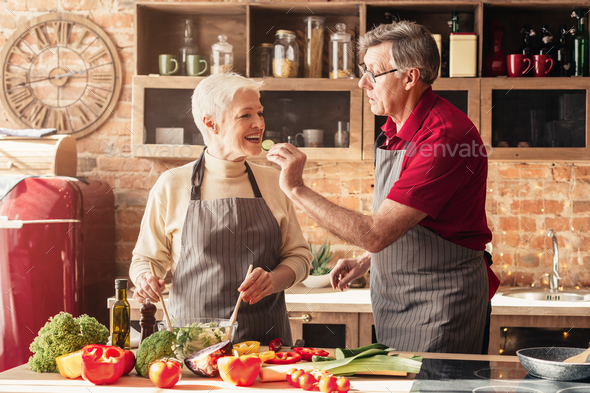 Romantic aged couple tasting food while cooking lunch in kitchen together