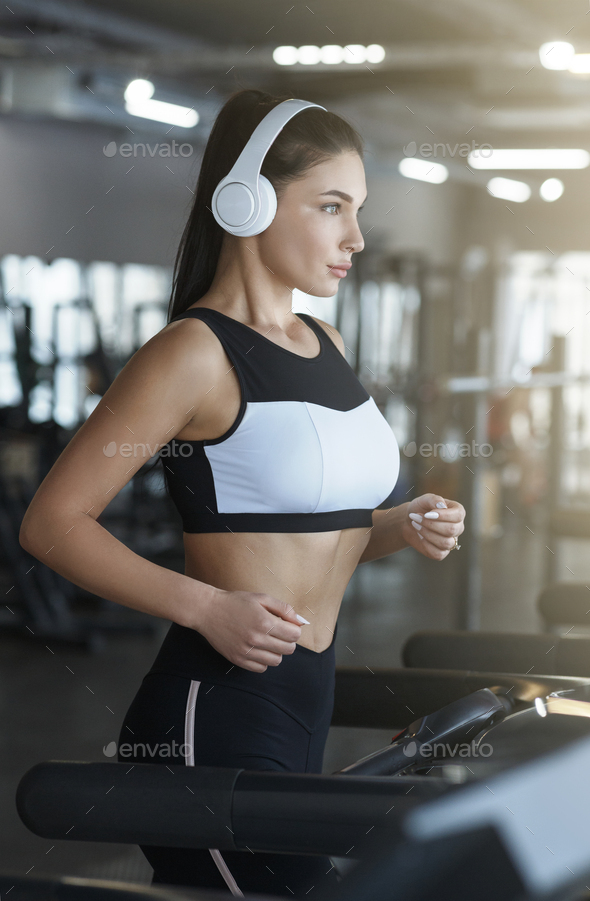 Sports workout. Young woman with headphones using treadmill in gym Stock Photo by Prostock-studio