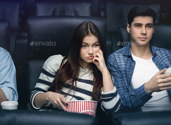 Boring date. Weary young girl with boyfriend watching movie in dark cinema
