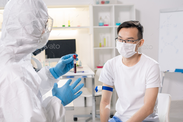 Sick chinese guy in protective mask sitting in front of lab worker with flask