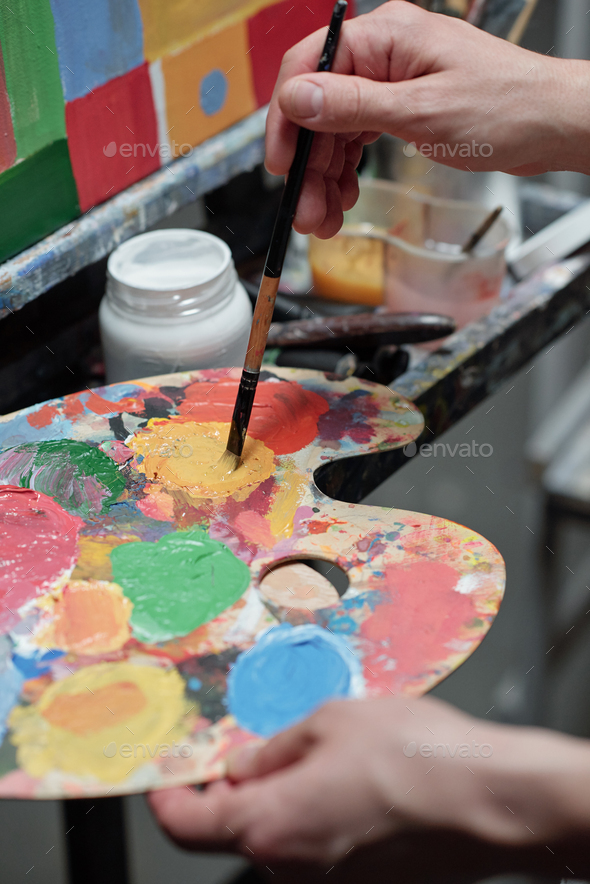 Hand of young professional painter mixing colors on palette in front of easel - Stock Photo - Images
