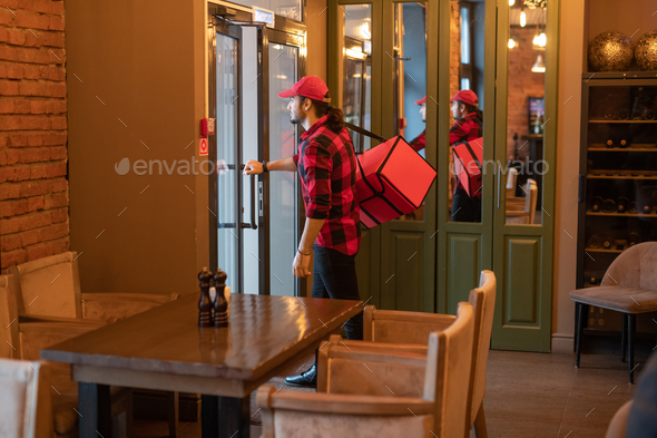 Young coutier with big red bag on shoulder going to open the door of cafe