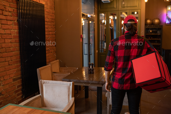 Rear view of young delivery service courier in flannel carrying big red bag
