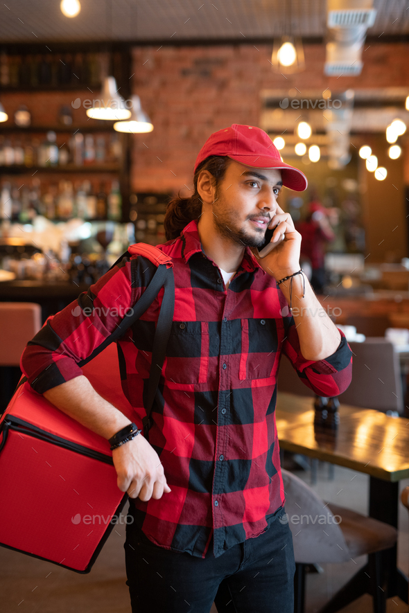 Young courier with big red bag on shoulder calling one of clients in cafe