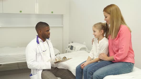 Cheerful Afro American Male Doctor Listen Consult Cute Caucasian Girl Make Notes in Patient Card at