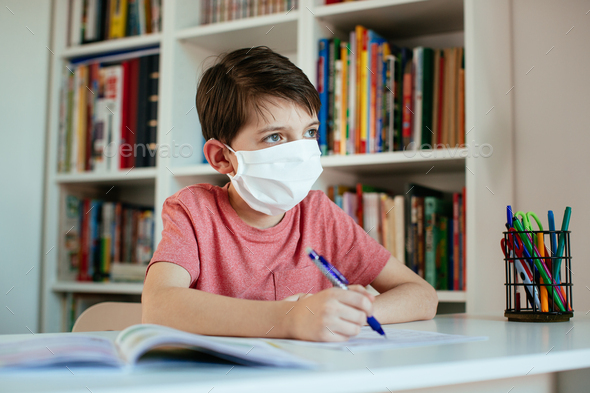 Young student wearing surgical mask working on school assignments on his own.