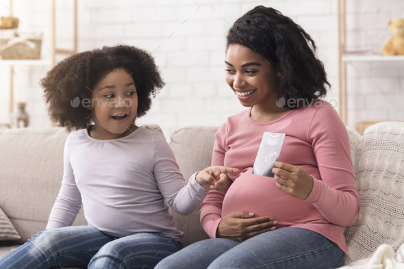 Pregnant afro mom showing ultrasound scan photo to her little daughter