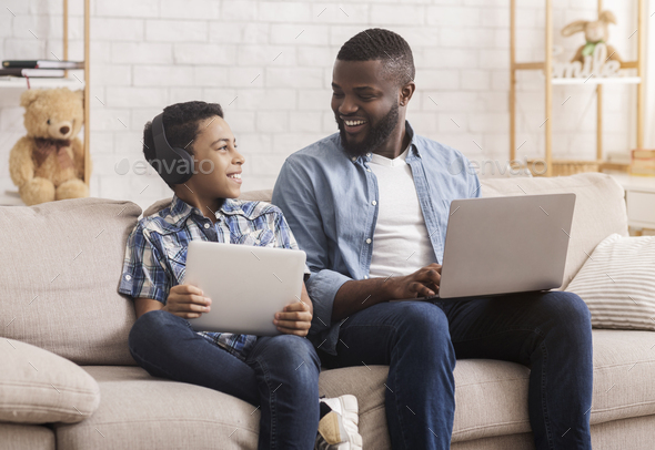 Black Father And Son Using Digital Tablet And Laptop At Home