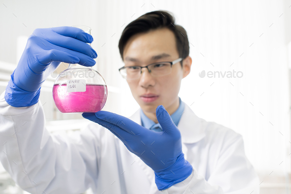 Young Chinese man in whitecoat and protective gloves studying chemical substance