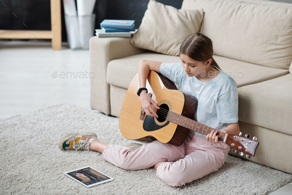 Pretty girl sitting on the floor by couch and learning to play guitar