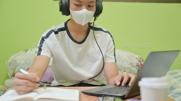 Male student with mask using a laptop and taking notes for exam preparation,He studied at home.