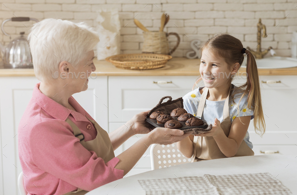Smiling girl and her granny holding tray with fresh chocolate muffins at home